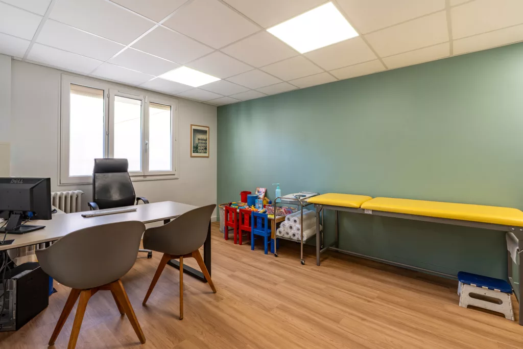 Grand cabinet médical aménagé pour la pédiatrie avec table d'examen jaune, bureau et espace de jeux pour enfants.