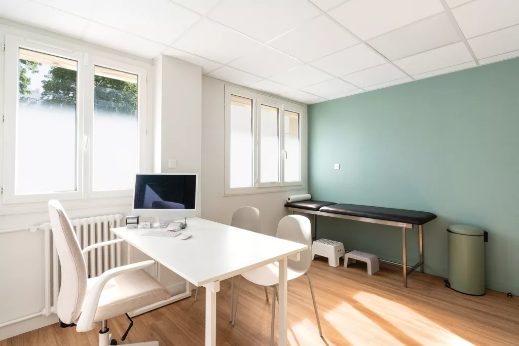 Salle d'examen médical lumineuse avec table d'auscultation, bureau de praticien et point d'eau sanitaire.