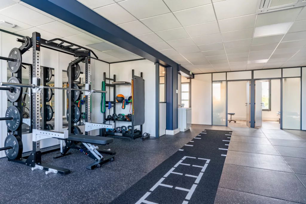 Zone de musculation équipée d'une cage de squat séparée des bureaux par des cloisons vitrées bord à bord.