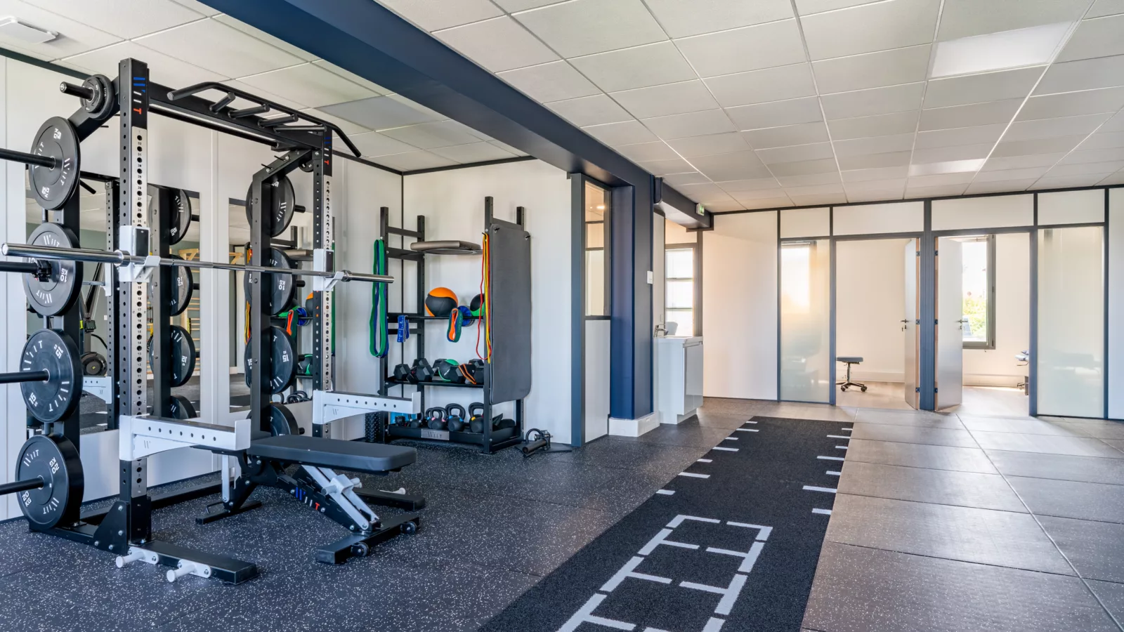 Zone de musculation équipée d'une cage de squat séparée des bureaux par des cloisons vitrées bord à bord.