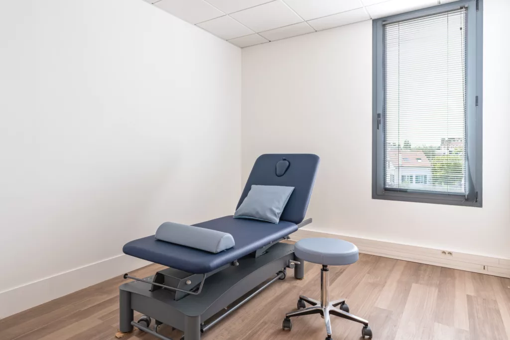 Salle de soin médicale lumineuse équipée d'une table d'examen avec stores vénitiens et sol PVC hygiénique.